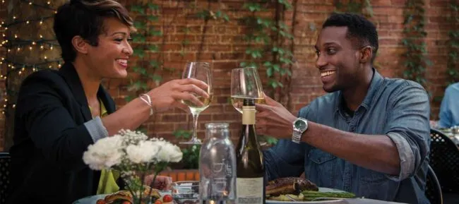 A smiling man and woman sit at an outdoor restaurant table, clinking wine glasses. Plates of food, a wine bottle, and a vase of white flowers are on the table. String lights and brick wall are in the background.