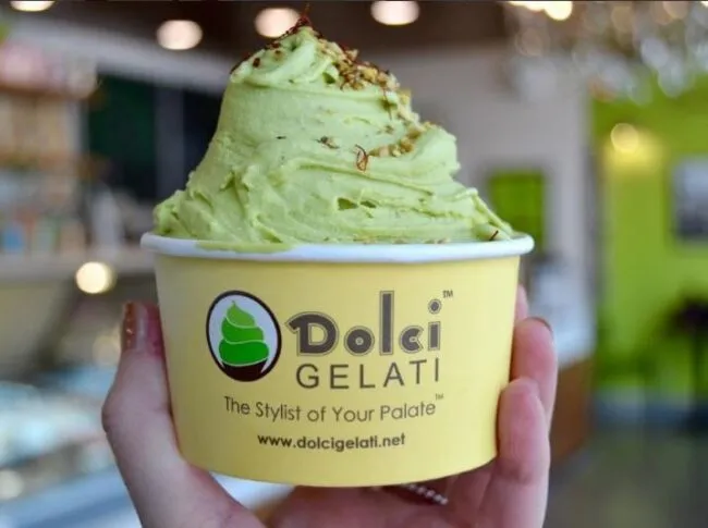 A hand holds a yellow cup labeled Dolci Gelati filled with a swirl of light green gelato, topped with a sprinkle of crushed nuts; a blurred ice cream shop background is visible.