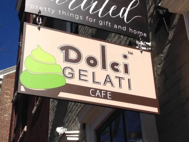 A shop sign for Dolci Gelati Cafe features the brand name in large letters and a green and brown swirl graphic resembling gelato. The sign hangs outside a brick building.