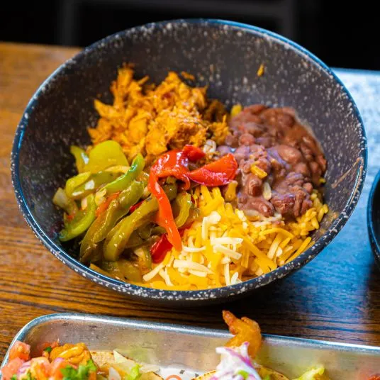 A black bowl filled with shredded chicken, sautéed peppers, refried beans, shredded cheese, and yellow rice, set on a wooden table.