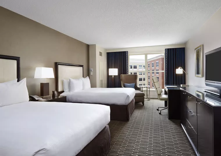 Hotel room with two queen beds, white bedding, nightstands with lamps, an armchair, desk with chair, TV, and large window overlooking nearby buildings. The room is modern and spacious with neutral colors.