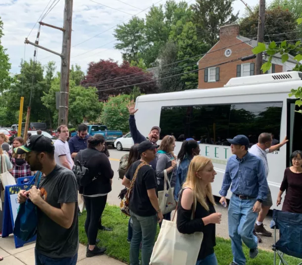 A group of people with tote bags gather and chat near a white shuttle bus parked on a suburban street lined with trees. One man in the middle waves and smiles at the camera.