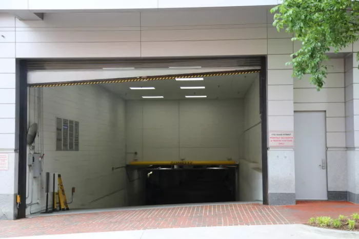 A large underground parking garage entrance with a partially open overhead door, a yellow clearance bar, and a brick sidewalk in front. Green tree branches are visible in the upper right corner.