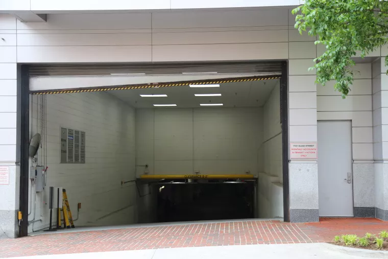 A large underground parking garage entrance with a partially open overhead door, a yellow clearance bar, and a brick sidewalk in front. Green tree branches are visible in the upper right corner.