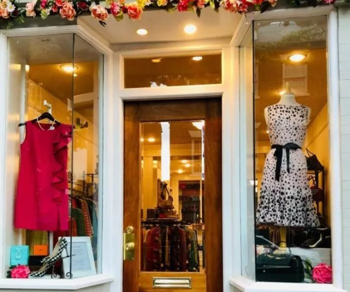 A clothing boutique storefront with a wooden door, mannequins displaying a pink dress and a white dress with black spots in glass windows, and a floral arrangement above the entrance.