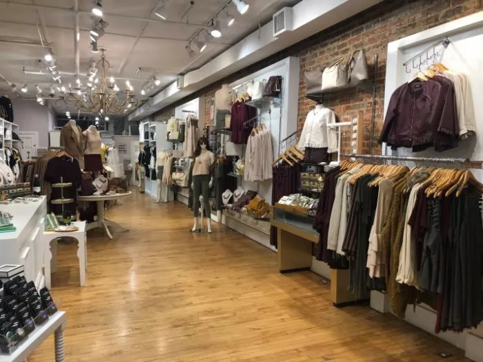 A boutique clothing store with wooden floors, exposed brick wall, and racks of neatly arranged womens clothes, bags, and accessories. Mannequins display outfits along the well-lit aisle.