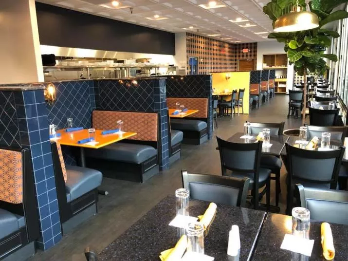 Modern restaurant interior with blue and orange booths on the left, black tables and chairs set with glasses and napkins on the right, and large windows letting in natural light.