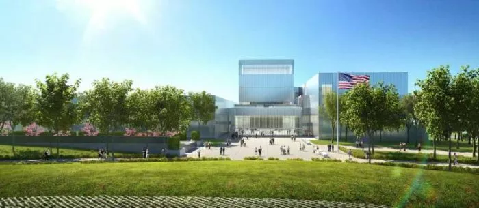 A modern glass building with multiple rectangular sections stands behind a spacious lawn, surrounded by green trees. People are gathered near the entrance, and an American flag is visible on the right side of the building.