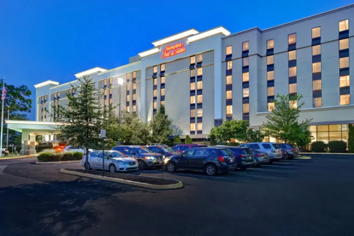 A well-lit Hampton Inn & Suites hotel at dusk, with many cars parked in the parking lot and trees surrounding the entrance. The building has multiple floors and illuminated windows.