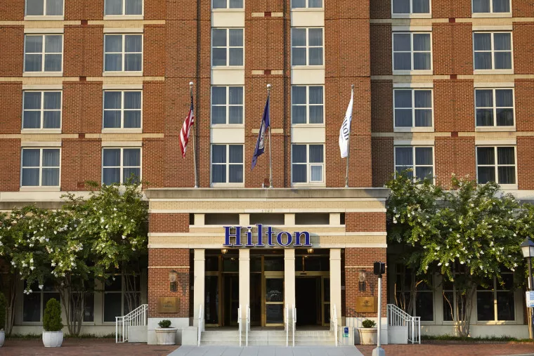 The front entrance of a Hilton hotel with a brick facade, three flags above the doorway, and greenery on either side of the entrance.