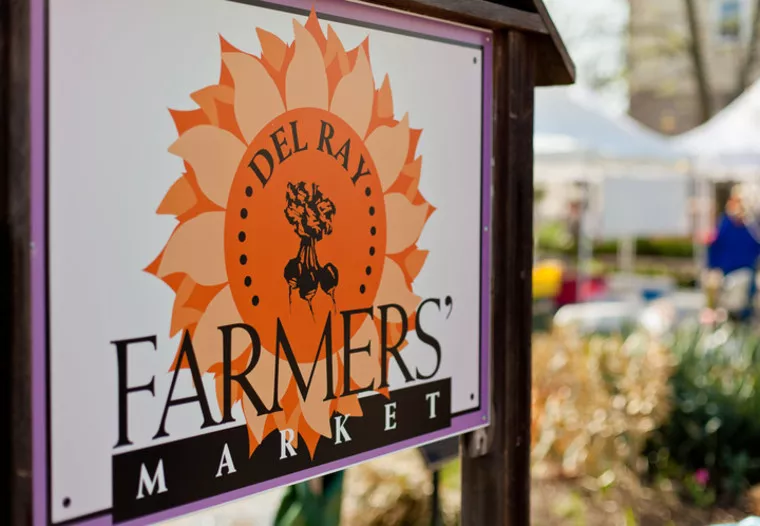 A sign reads Del Ray Farmers Market with a sunburst design and tree illustration; blurred tents and greenery from the market are visible in the background.
