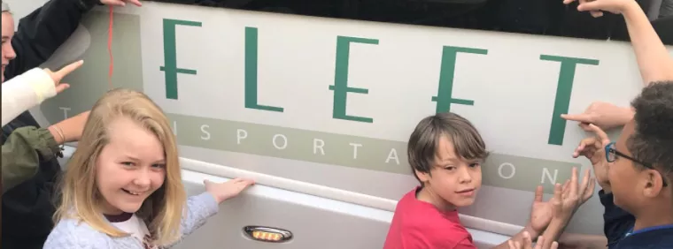 A group of smiling children stand in front of a vehicle, touching and pointing at a sign that reads FLEET TRANSPORTATION.