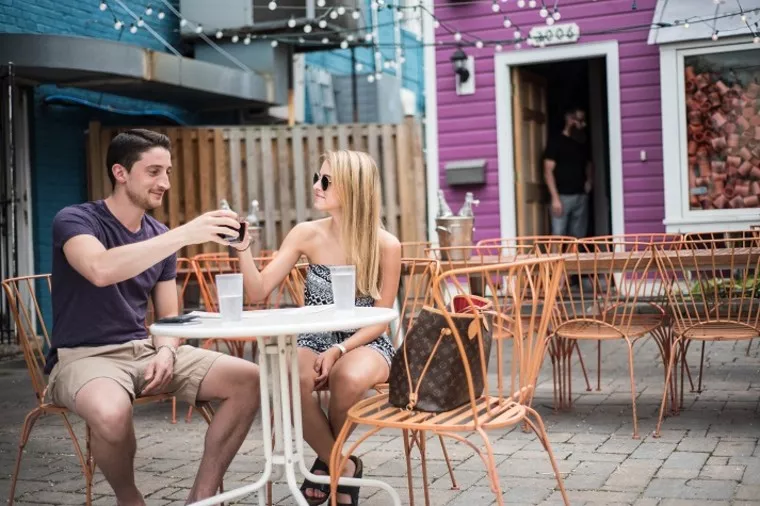 A man and woman sit at an outdoor café table, toasting drinks and smiling. Orange metal chairs surround them, and string lights hang above. A man stands in the doorway of a bright purple building in the background.