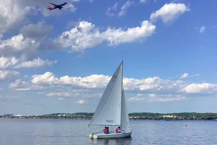 A sailboat with two people glides on calm water under a partly cloudy sky, while a commercial airplane flies overhead. In the background, there is a shoreline with buildings and greenery.