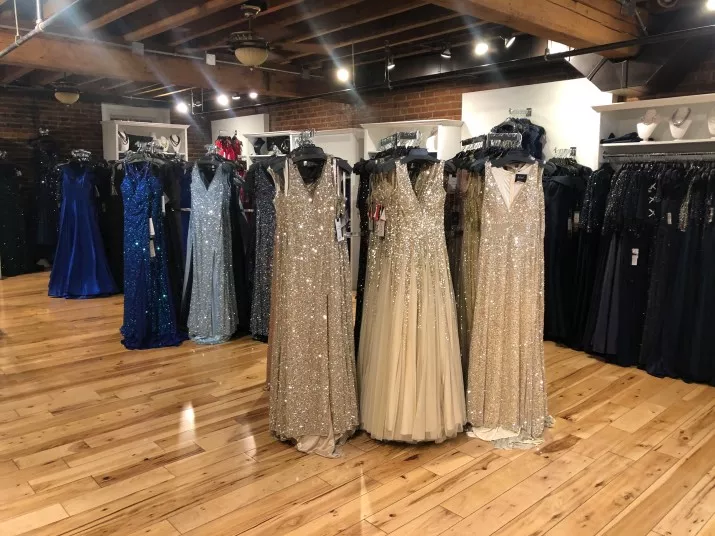 A boutique showroom with shiny wooden floors displays elegant floor-length evening gowns, including three sparkling gold dresses in the center and various blue and black dresses on racks behind them.