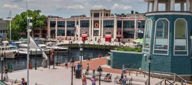People walk along a waterfront boardwalk with docked boats, a teal building with a cupola, and a large building with red awnings in the background under a partly cloudy sky.
