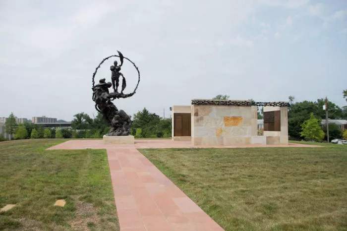 A large outdoor bronze sculpture of soldiers in action stands next to a stone memorial structure on a grassy area, with a red tiled walkway leading to both. Trees and buildings are visible in the background under a cloudy sky.