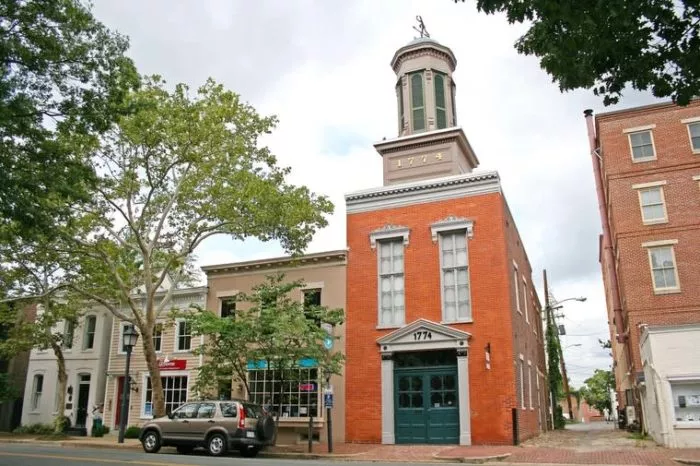 Historic red-brick firehouse with “1774” above the arched garage door, located on a tree-lined street with parked cars and neighboring storefronts on a cloudy day.