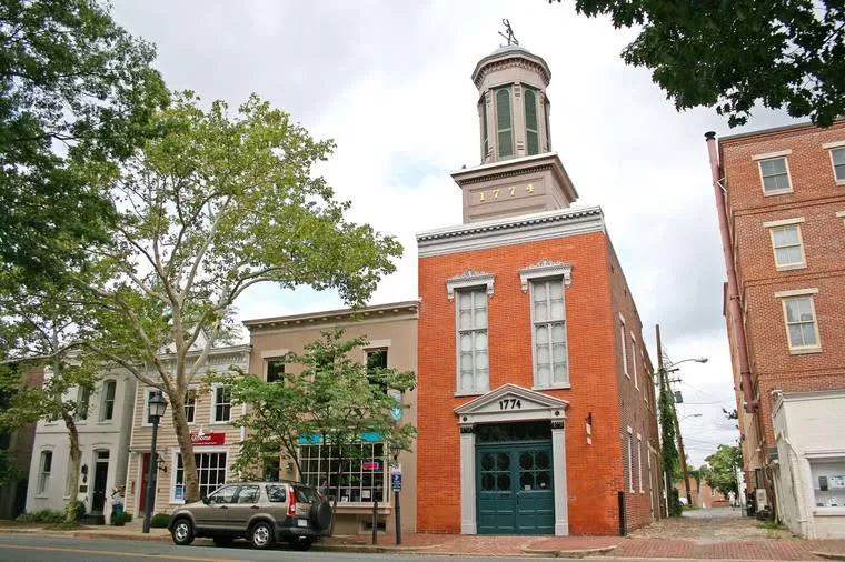 Historic red-brick firehouse with “1774” above the arched garage door, located on a tree-lined street with parked cars and neighboring storefronts on a cloudy day.