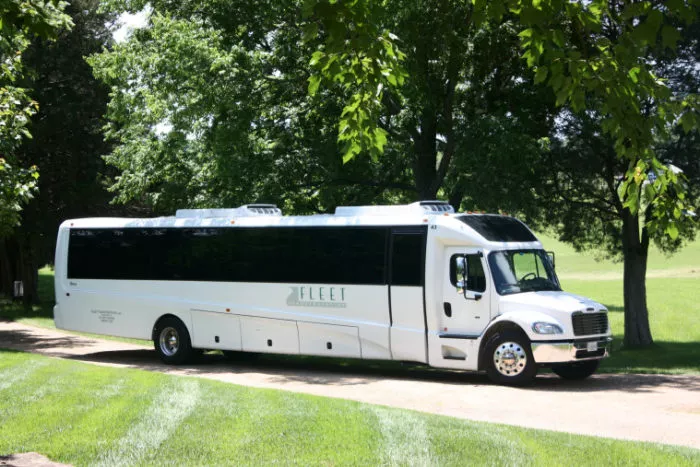 A large white tour bus labeled Fleet is parked on a paved road surrounded by green grass and trees on a sunny day.