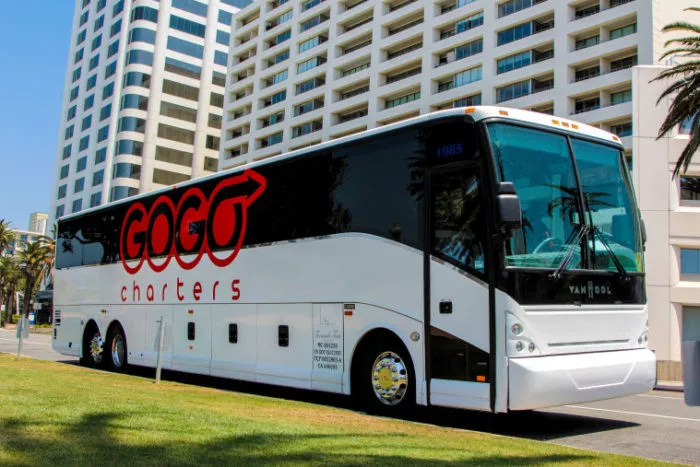 A large white charter bus with GOGO charters written on the side is parked on a street in front of a tall, modern hotel building with palm trees nearby.