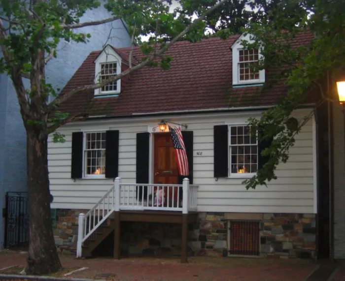 A small white house with black shutters, a red shingle roof, stone foundation, front porch, and steps. An American flag hangs by the door. The house is partially shaded by a large tree.