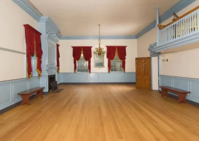 Spacious, empty room with light wood floors, pale blue trim, red curtains, a chandelier, a fireplace, two wooden benches, a wardrobe, and an upper balcony with a railing.