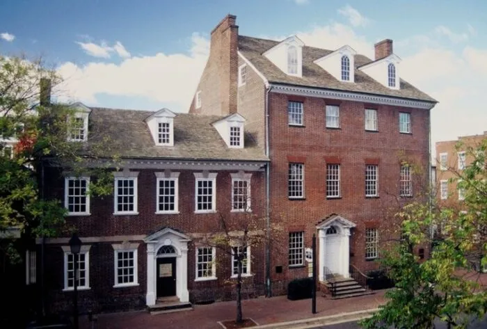 A large, historic red-brick building with white trim, tall windows, dormers, and two arched doorways, sits along a tree-lined street under a blue sky with scattered clouds.