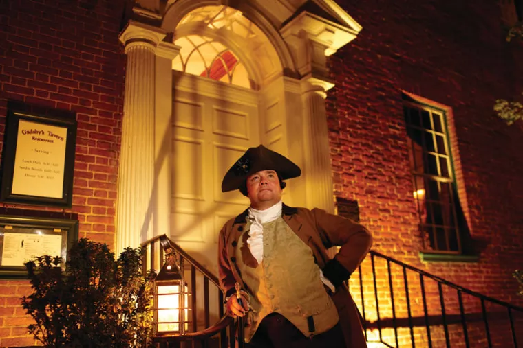 A person in colonial-era clothing stands confidently on brick steps outside a historic building at night, lit warmly by a lantern. A sign by the door reads “Gadsby’s Tavern.”.