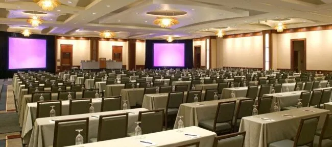 A large, well-lit conference room with rows of tables and chairs facing two large projection screens. Each table is set with a notepad, pen, and water glass, ready for a presentation or seminar.