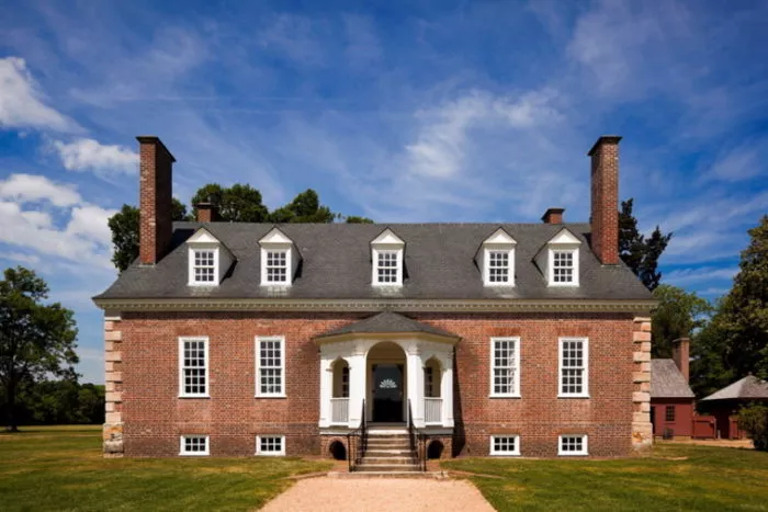 A symmetrical, red-brick colonial-style house with white trim, dormer windows, and two large chimneys, set against a blue sky with scattered clouds and surrounded by green grass and trees.