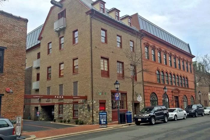 A brick parking garage with red-trimmed windows and an entrance marked “PARK” stands on a street lined with parked cars and historic-style buildings under a cloudy sky.