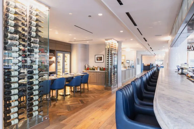 Modern restaurant interior with a curved marble bar and blue leather stools, a wine display cabinet, wood flooring, and a dining area with blue chairs and elegant table settings. Bright and spacious atmosphere.