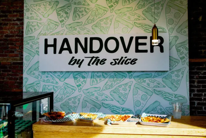 A sign reading HANDOVER by The slice is displayed above a counter with trays of pizza slices. The background features drawings of pizza slices, and a single hanging light shines on the sign.