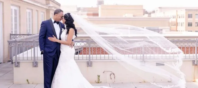 A bride and groom embrace on a rooftop. The bride wears a long white gown and veil flowing in the wind; the groom is in a navy blue suit. They look lovingly at each other against an urban cityscape background.