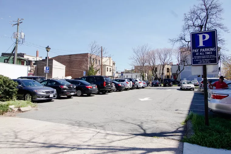 A small parking lot filled with cars on a sunny day, with a blue sign that reads “Pay Parking at All Times.” Buildings and bare trees are visible in the background.