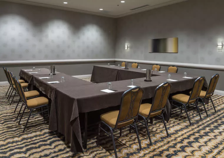 A conference room with brown tables arranged in a U-shape, surrounded by chairs. Each table has a water pitcher and glasses. The room has patterned carpet, gray wallpaper, and soft overhead lighting.