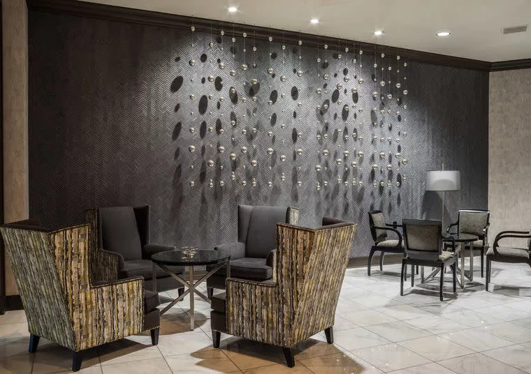 Modern lounge area with patterned armchairs, a glass coffee table, dark accent wall featuring decorative hanging elements, and a small dining table with chairs in the background, all set on a tiled floor.