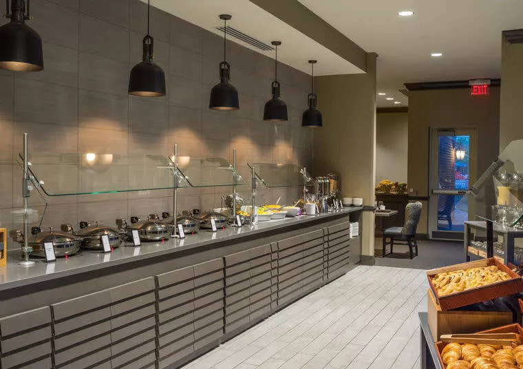 A modern buffet area with various breakfast items in chafing dishes, overhead black pendant lights, and trays of pastries on the side, in a clean, well-lit dining space.