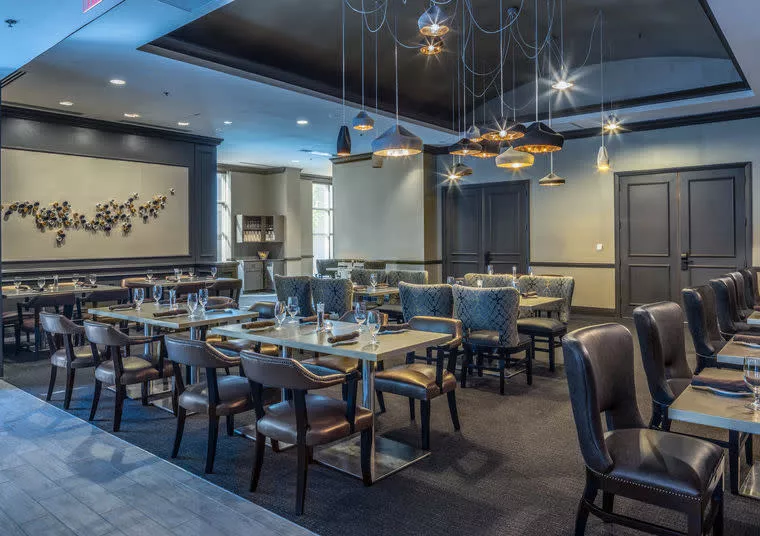 Modern restaurant dining area with elegant decor, featuring cushioned chairs, set tables with glassware, overhead pendant lighting, and wall art in a neutral color palette. The space appears clean and inviting.
