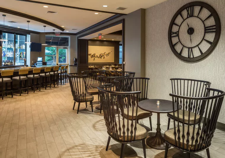 Modern lounge area with wooden floor, large wall clock, black spindle-back chairs around small tables, and a bar with high stools and shelves of bottles in the background. Warm lighting and contemporary decor.
