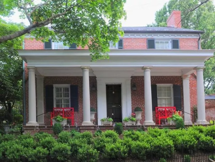 A two-story brick house with green shutters, white columns, and a wide front porch. Red chairs and potted plants decorate the porch, and a neatly trimmed hedge lines the front yard.