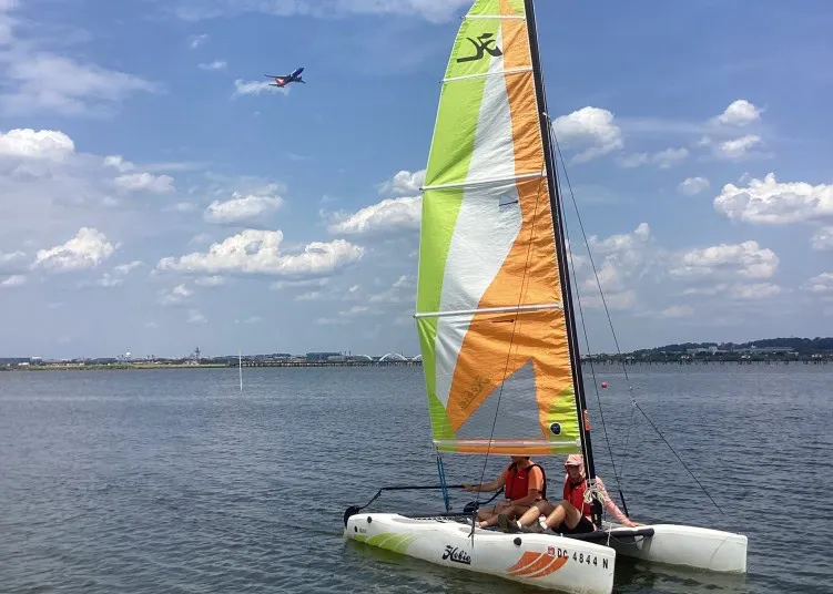 Two people in life jackets sail a small catamaran with colorful orange, green, and white sails on calm water. In the sky above, an airplane is flying, with a distant cityscape under a blue, partly cloudy sky.