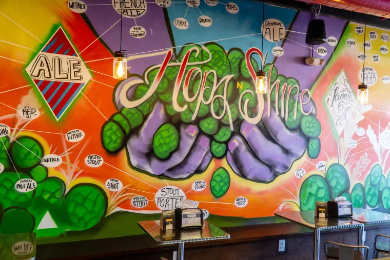 A colorful mural of large hands holding green hops with “Hop n Shine” written above, surrounded by names of beer styles on a vibrant wall in a restaurant with tables and hanging lights in front.
