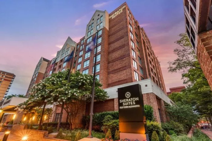 Exterior view of Sheraton Suites Old Town Alexandria hotel at sunset, showing a multi-story brick building with large windows, trees, and a prominent black and white sign in the foreground.