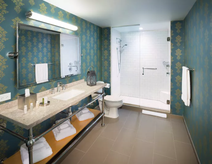 A stylish bathroom with teal-patterned wallpaper, a large mirror above a marble countertop, neatly folded towels on a lower shelf, a walk-in glass shower, and a toilet next to the vanity.