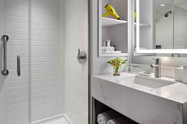 Modern bathroom with a glass-enclosed shower, white tiled walls, a marble countertop sink, a mirror, shelves holding a yellow hairdryer and towels, and a small vase with green flowers.