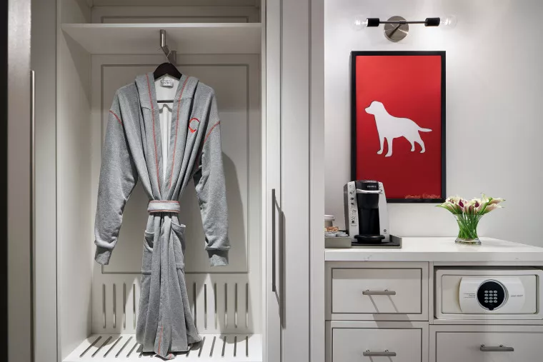 A gray bathrobe with red trim hangs in a built-in closet. Next to it, a countertop holds a coffee maker, vase of flowers, and a safe, with a framed red print of a dog on the wall above.