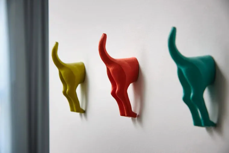 Three colorful wall hooks shaped like the back halves of dogs—one yellow, one red, and one teal—are mounted in a row on a white wall.