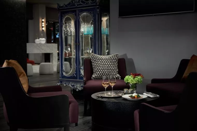 Four dark armchairs surround a round black table with wine glasses, a small plate of food, and a flower arrangement in a modern, dimly lit lounge. A decorative blue cabinet stands in the background.